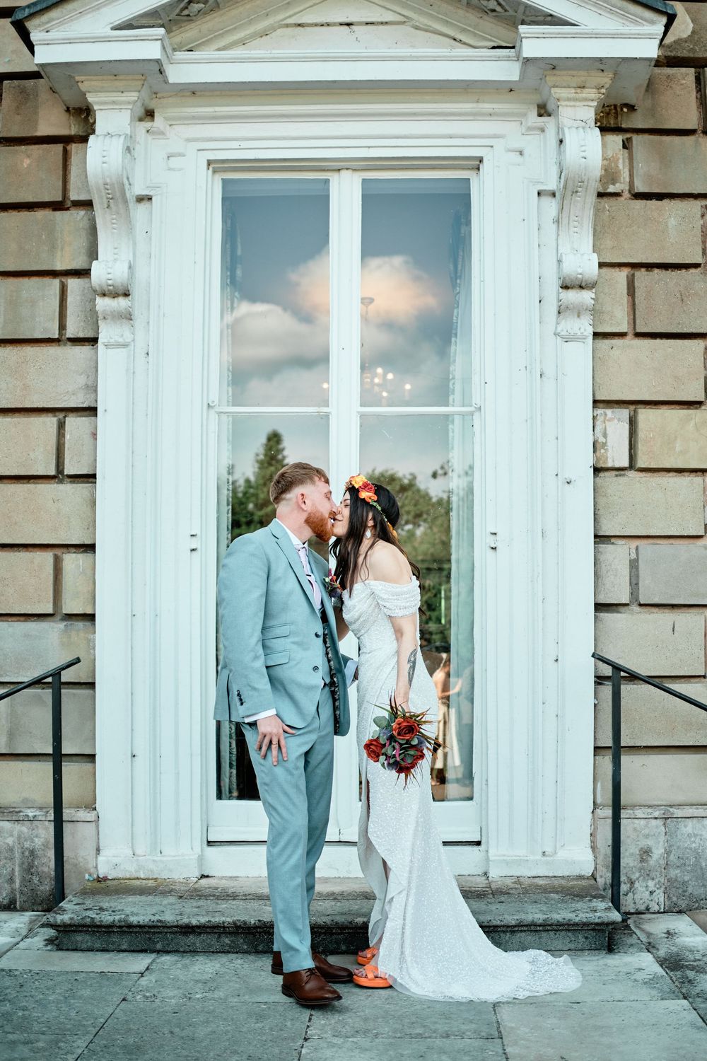 Elegant couple shares a romantic moment in front of a grand white doorway with cloud reflections in window panes.