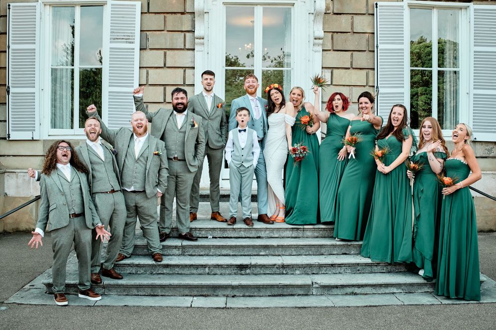 Wedding party in matching green dresses and gray suits poses on grand stone steps outside elegant building.