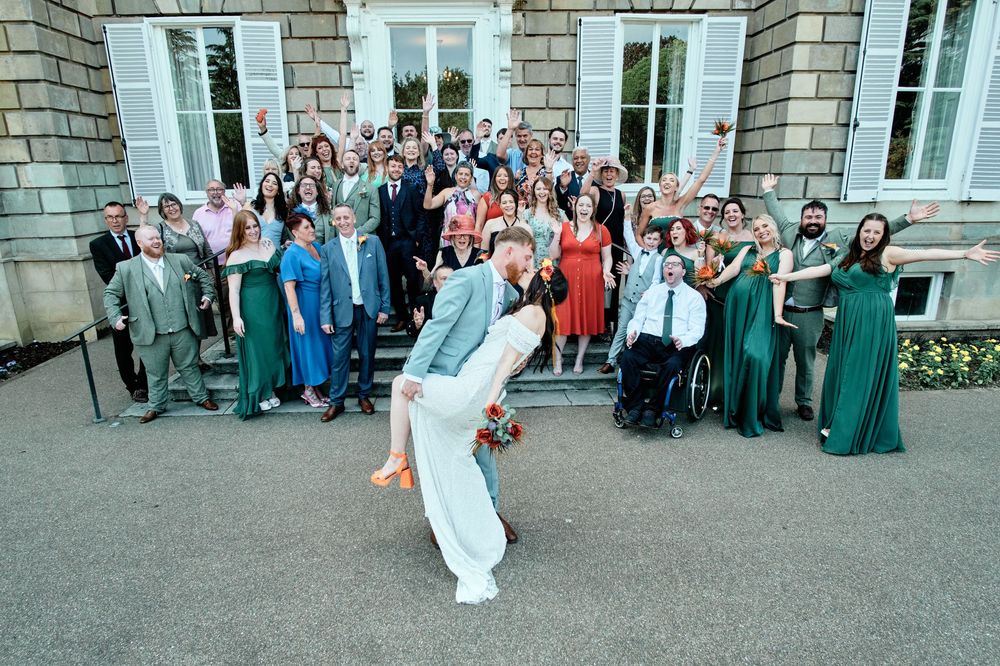 A line of wedding guests stand in a receiving line outside a grand stone building for a group photo.