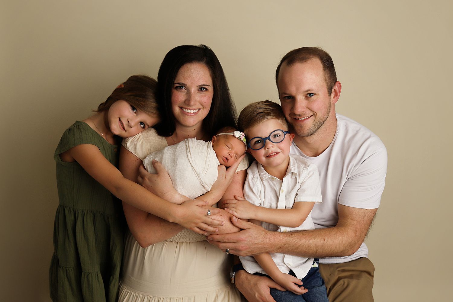 Family wearing white and olive clothing pose together in loving embrace against neutral beige background.
