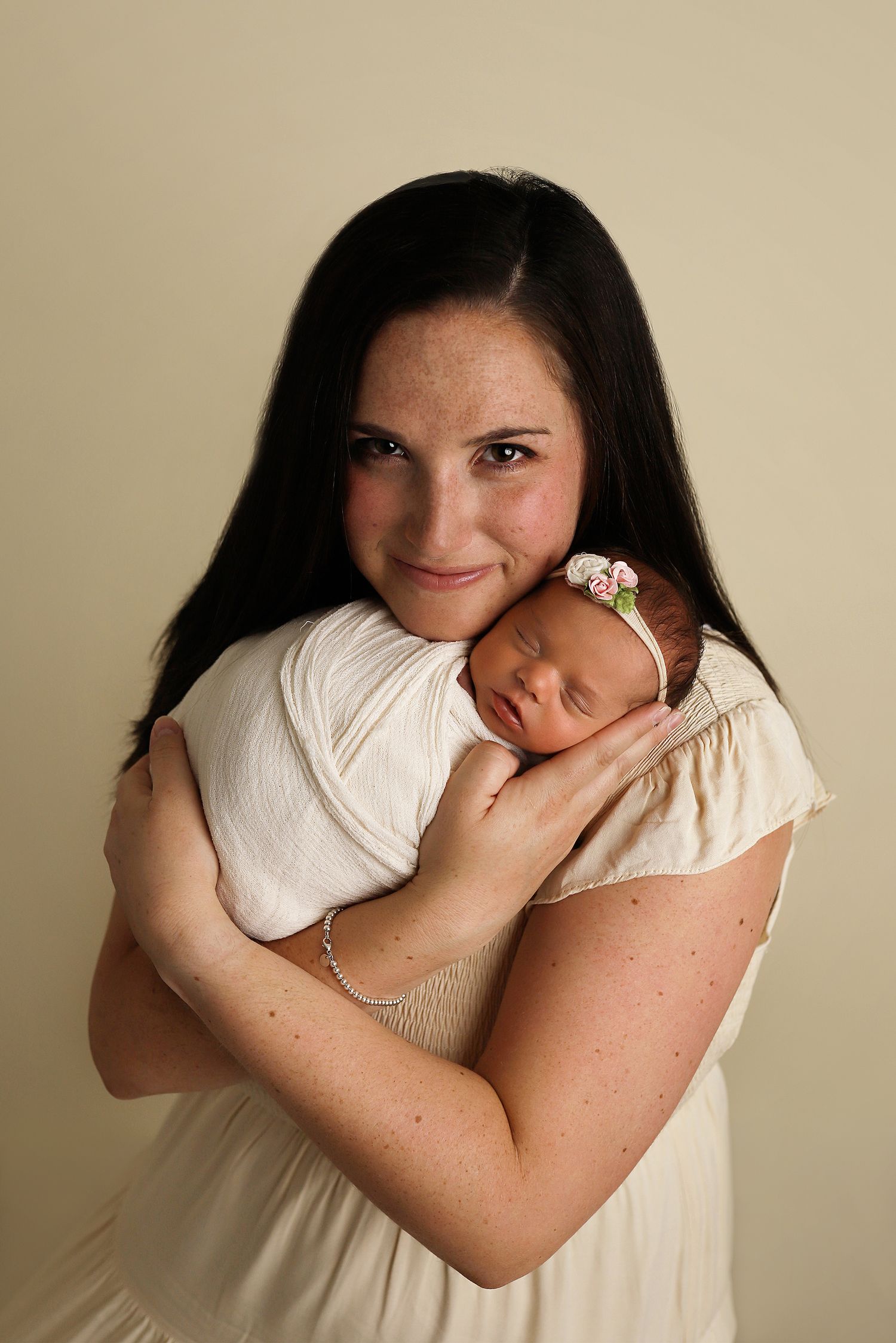 A mother in a cream-colored dress tenderly holds her sleeping newborn baby against her chest in a studio portrait.