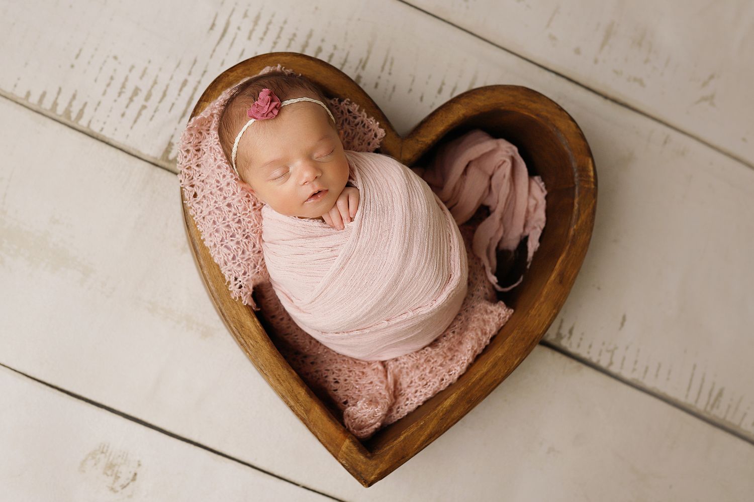Heart-shaped wooden prop with baby in pink wrap photographed from above in artistic newborn portrait style.