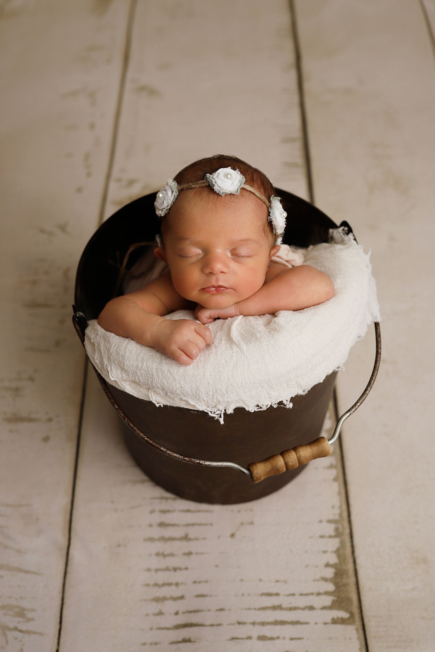 Sleeping newborn rests peacefully on white cushion in vintage metal bucket on wooden floor.