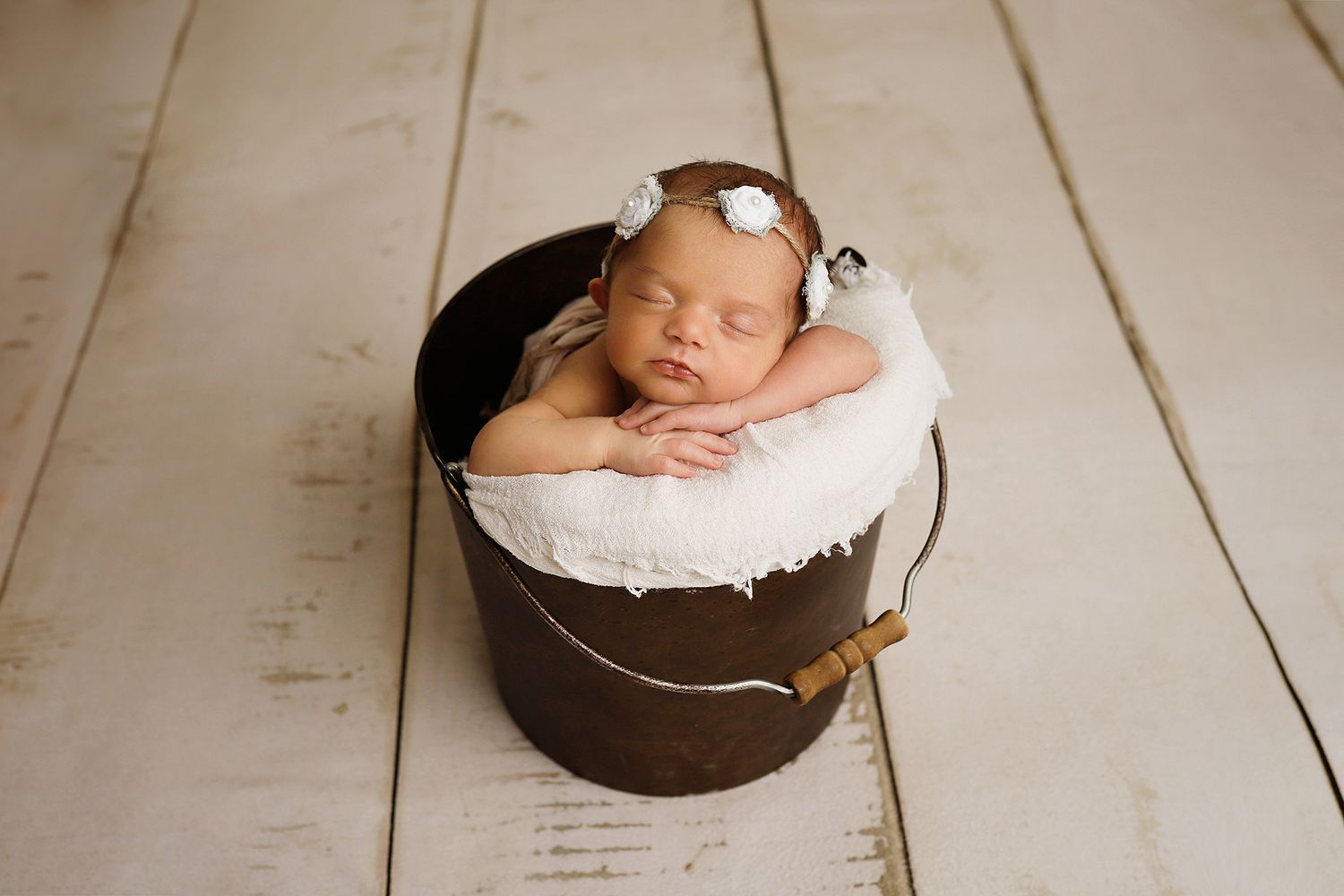 A sleeping newborn baby nestled in a dark bucket with white cushioning on a wooden floor.