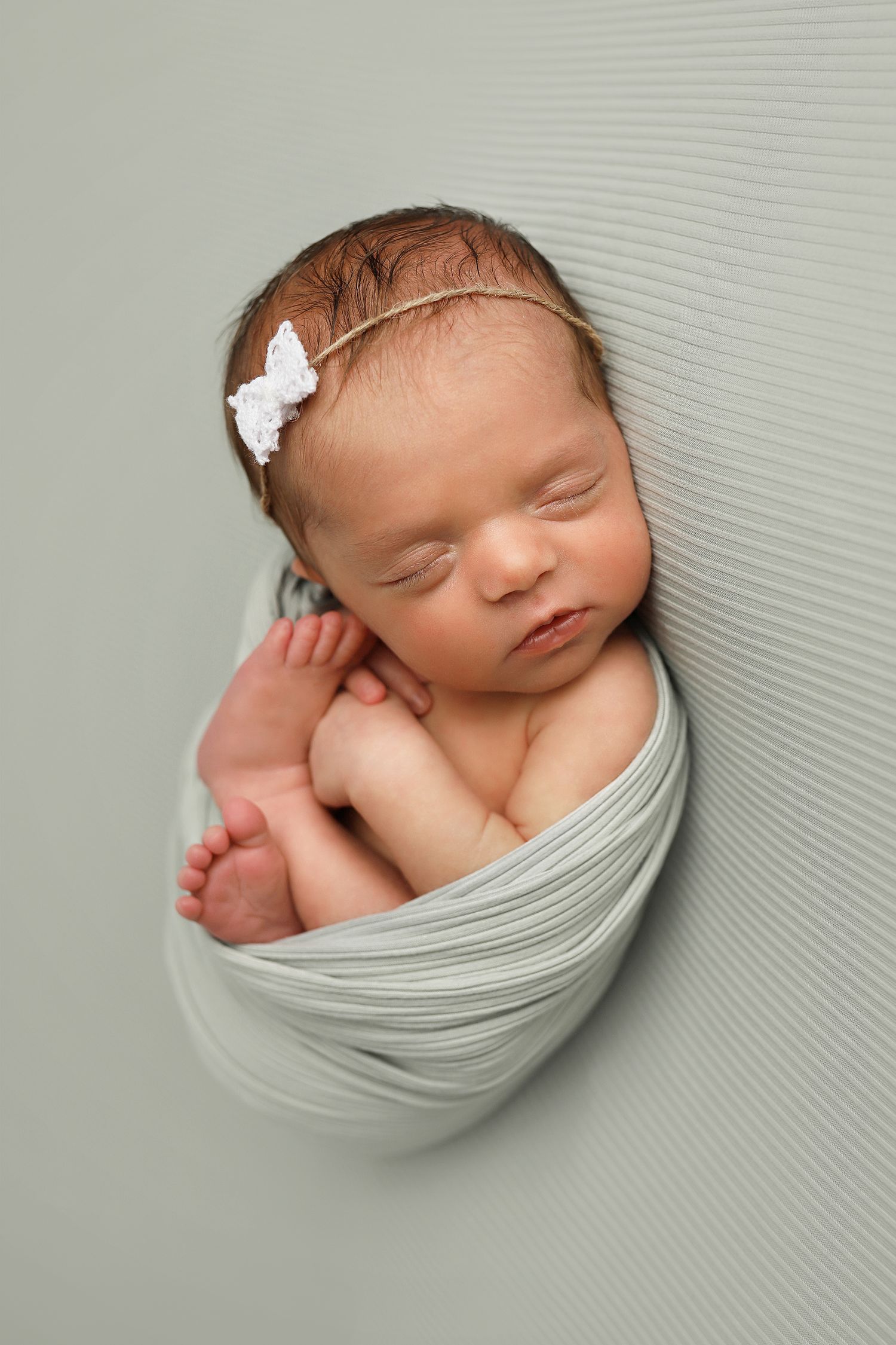 Sleeping newborn baby swaddled in soft gray fabric against a light background with tiny white hair accessories.