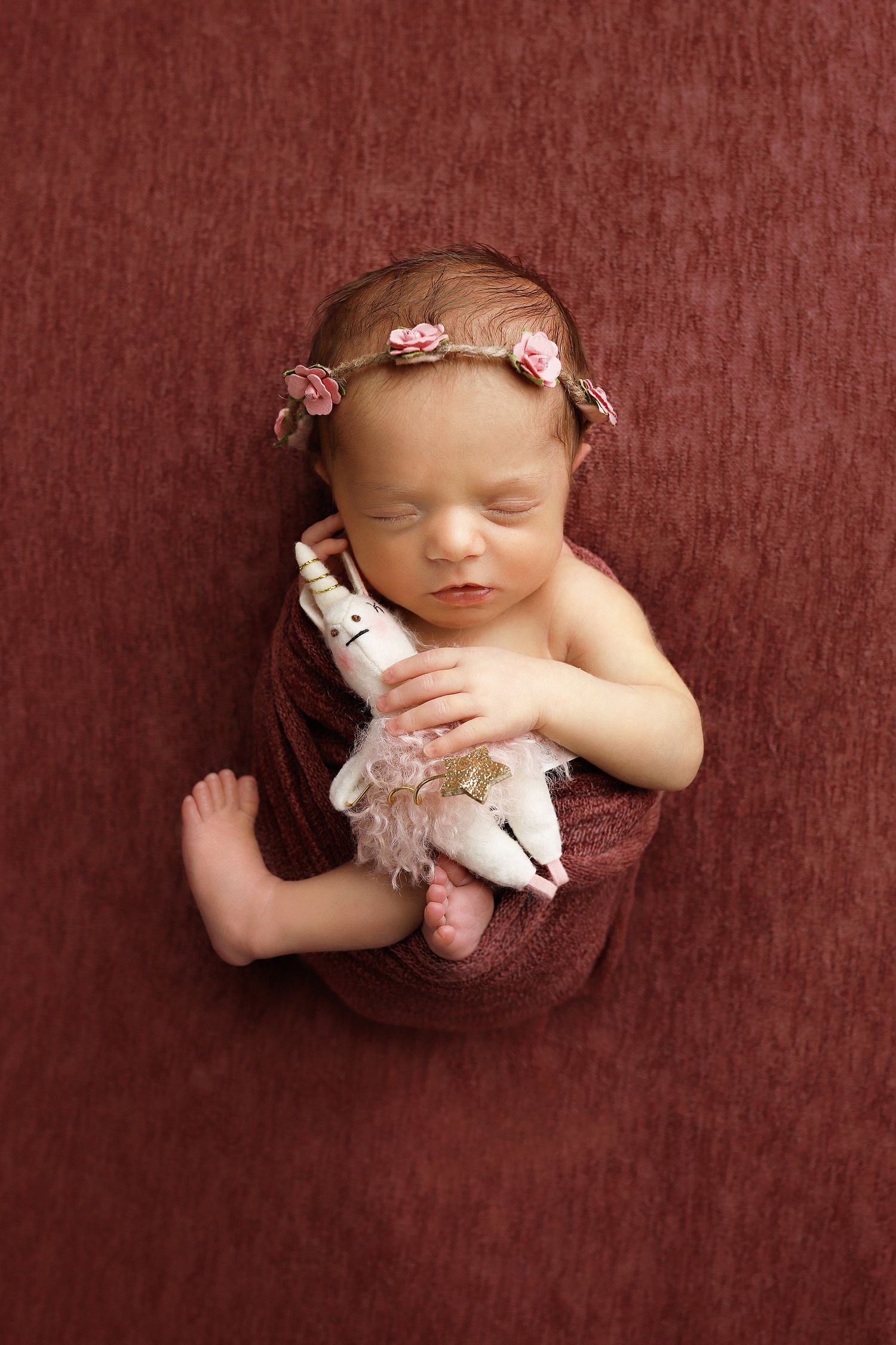 A sleeping newborn baby wrapped in brown fabric holding a small teddy bear against a dark red textured background.