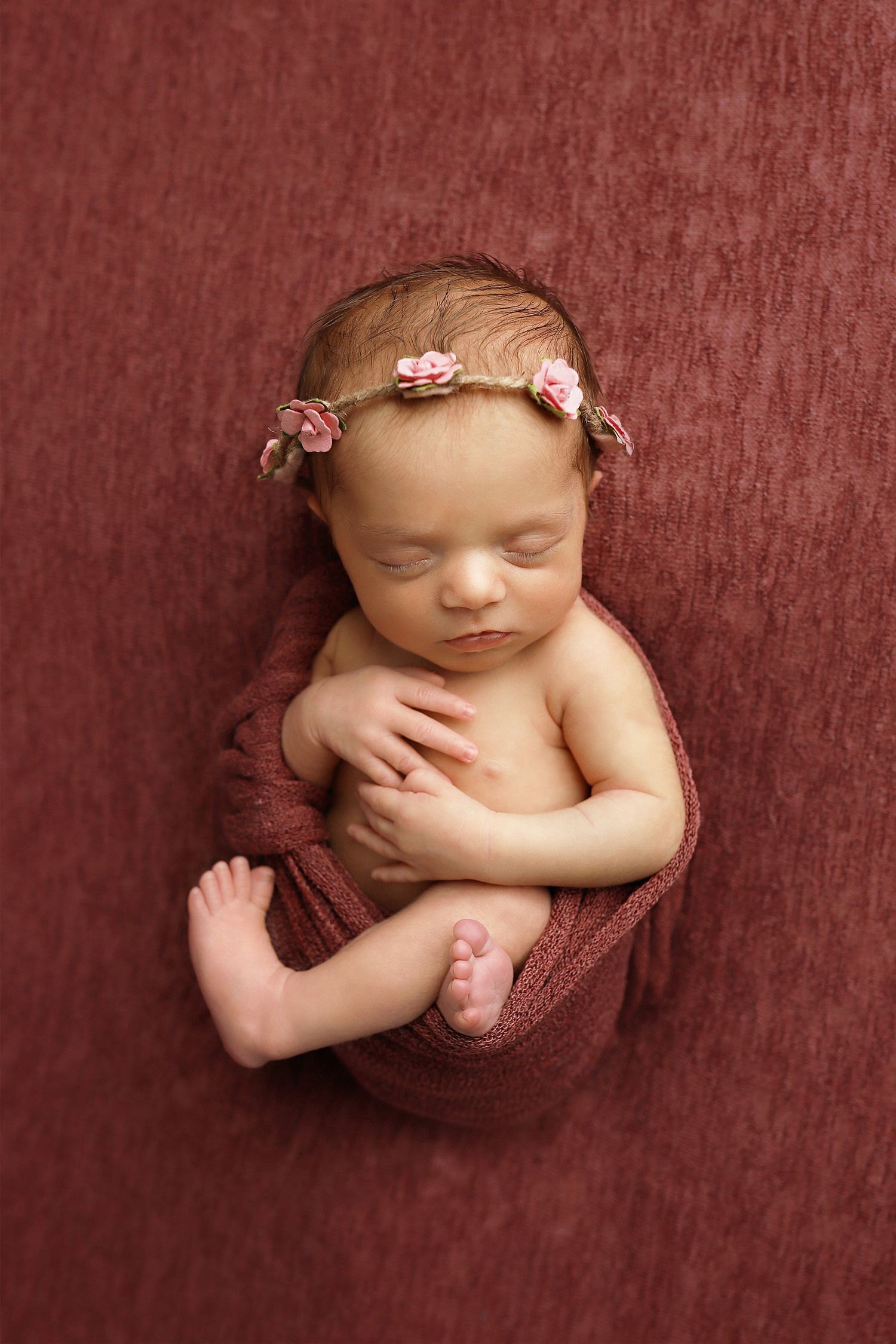Utah newborn photography Sleeping newborn baby with delicate pink floral headband curled up on burgundy textured fabric.