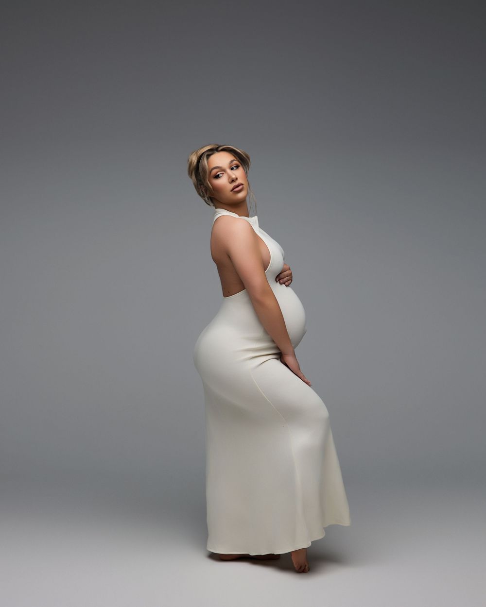 A figure in a long white form-fitting dress poses elegantly against a gray studio backdrop.