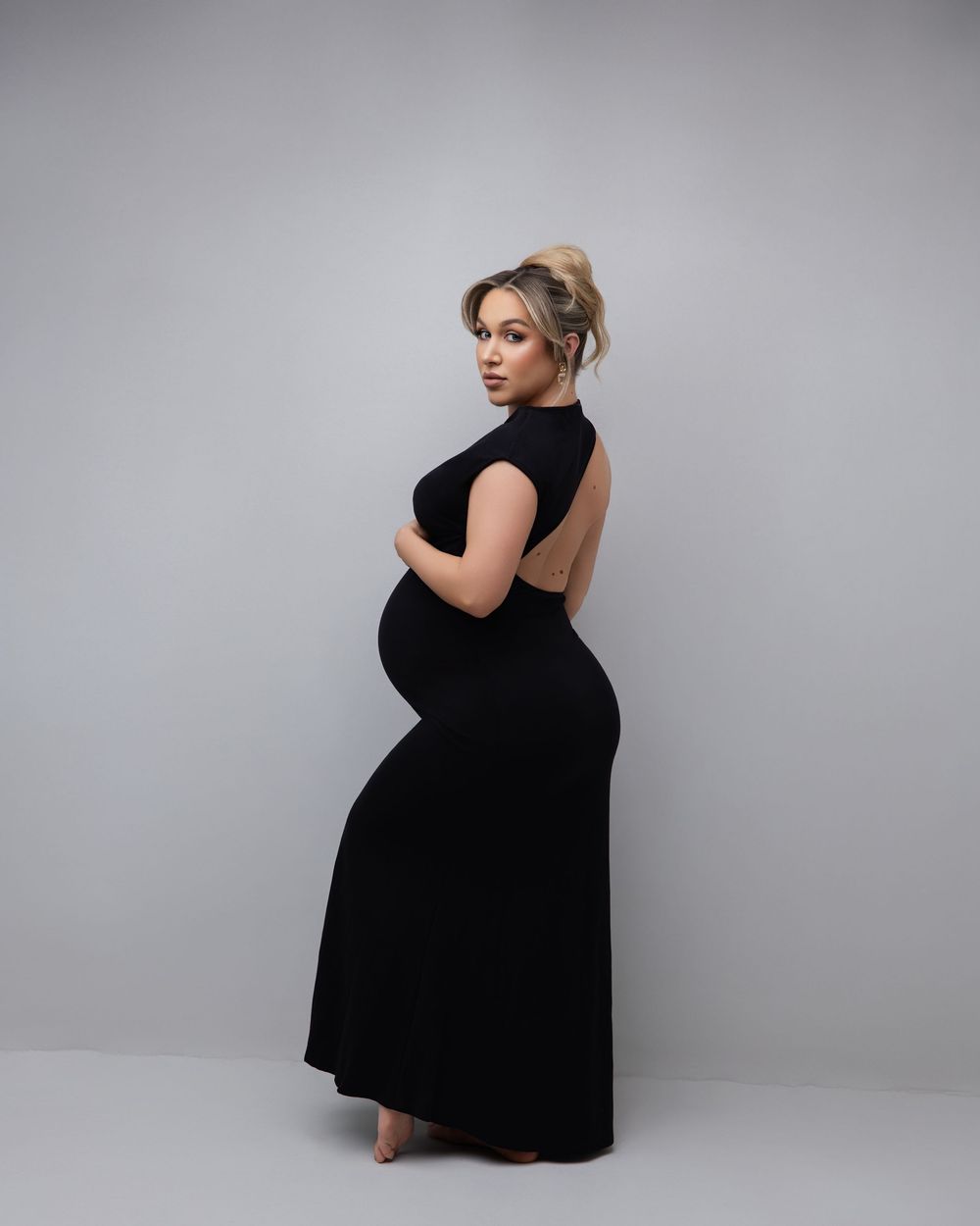 A pregnant woman in a long black dress poses elegantly against a gray studio backdrop.