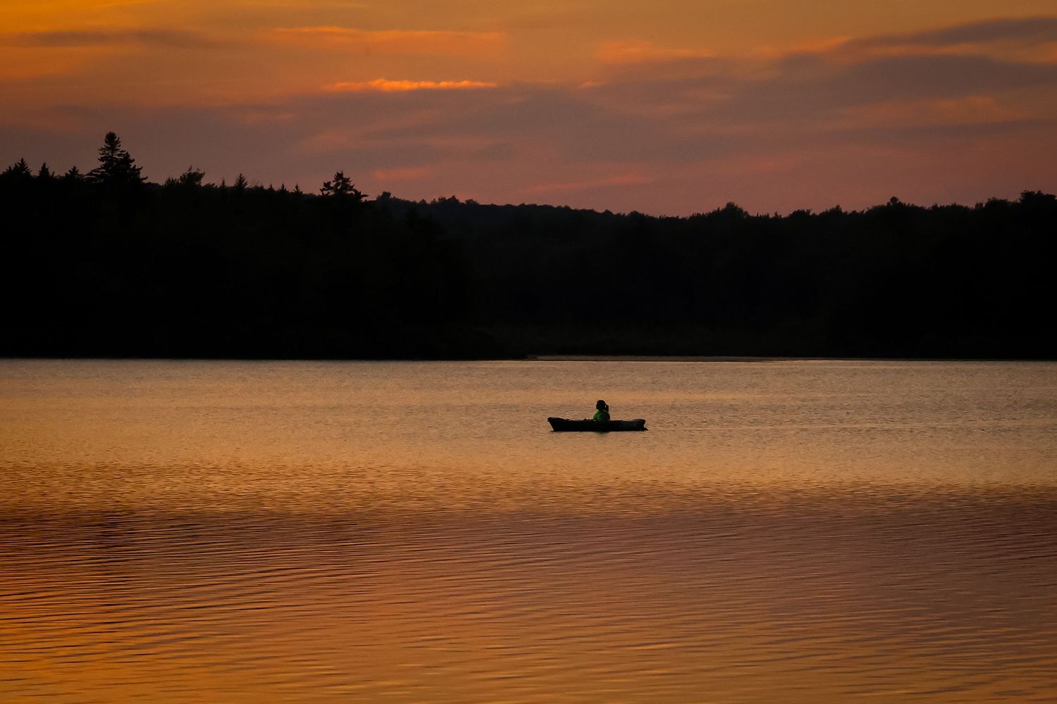 Silhouette of a small boat on calm lake waters during golden sunset with dark treeline on horizon.