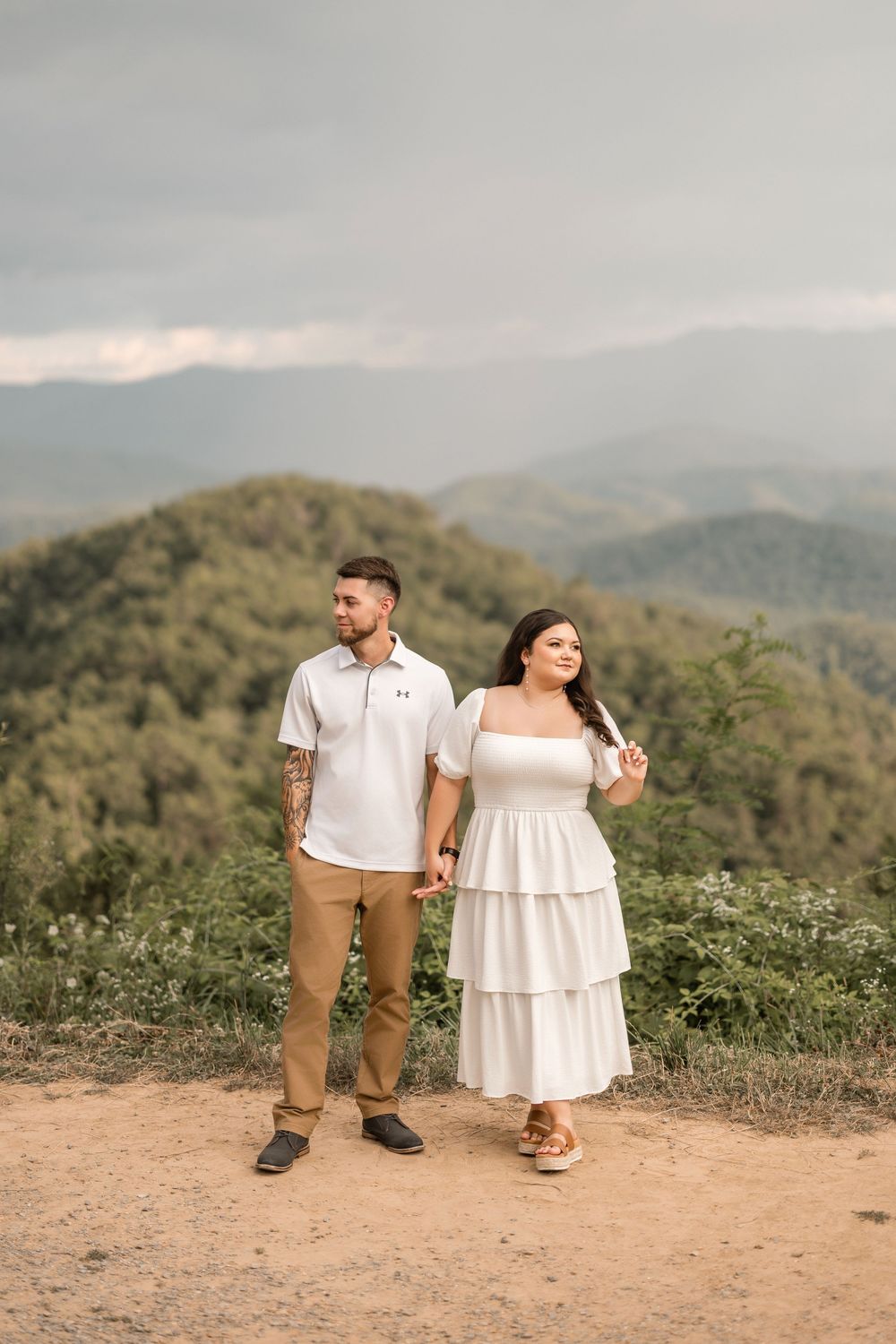 A couple stands on a mountain overlook together with scenic rolling hills and mountains in the background during sunset.