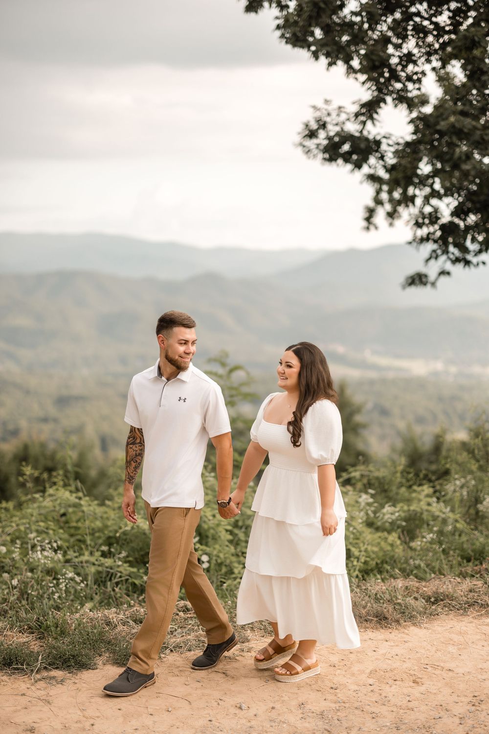 A couple holds hands while walking along a scenic mountain trail with rolling hills in the background at sunset.