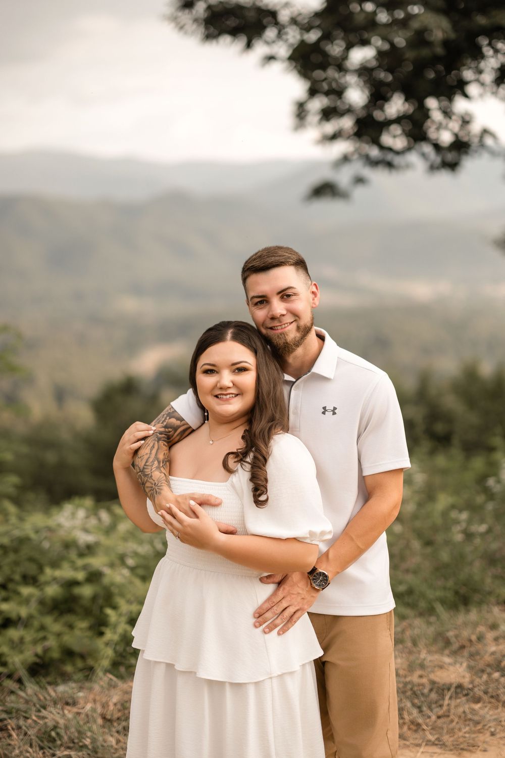 A couple embraces outdoors on a hilltop with mountains and trees visible in the background during golden hour.