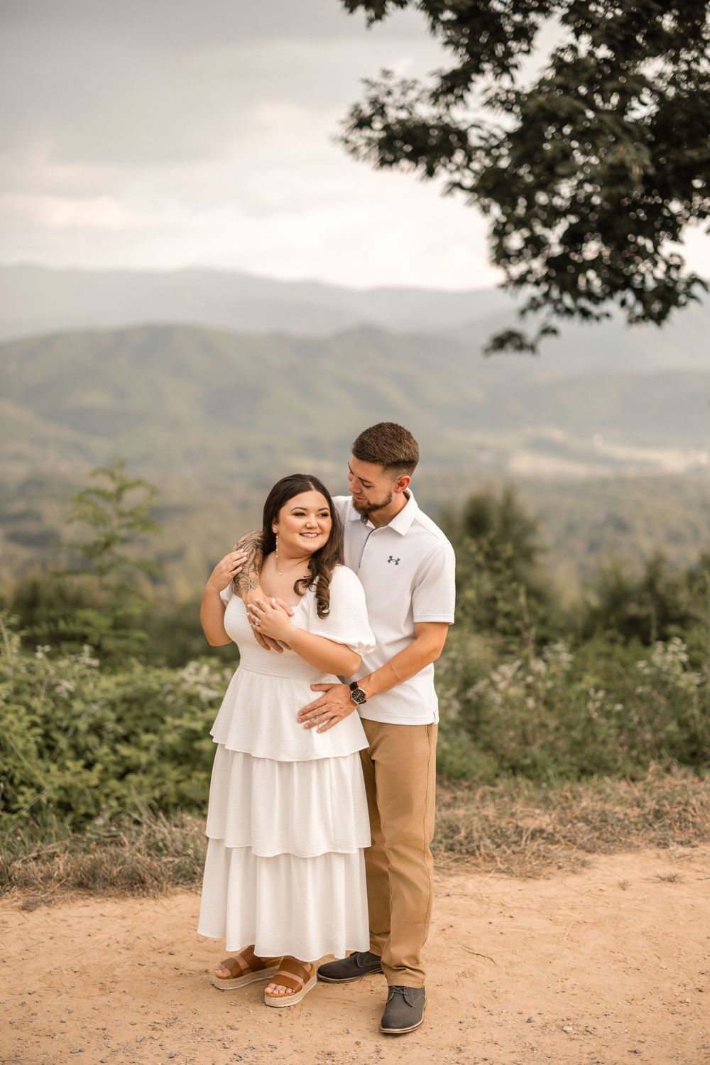 A couple embraces on a scenic mountaintop trail with rolling hills in the background during golden hour.