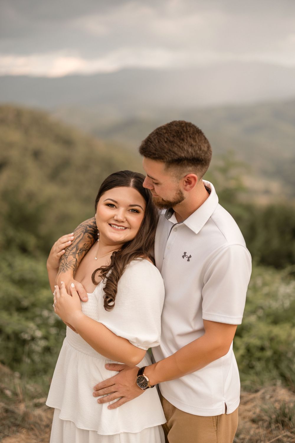 A couple in coordinating white outfits embrace lovingly in an outdoor mountain setting during a romantic photoshoot.
