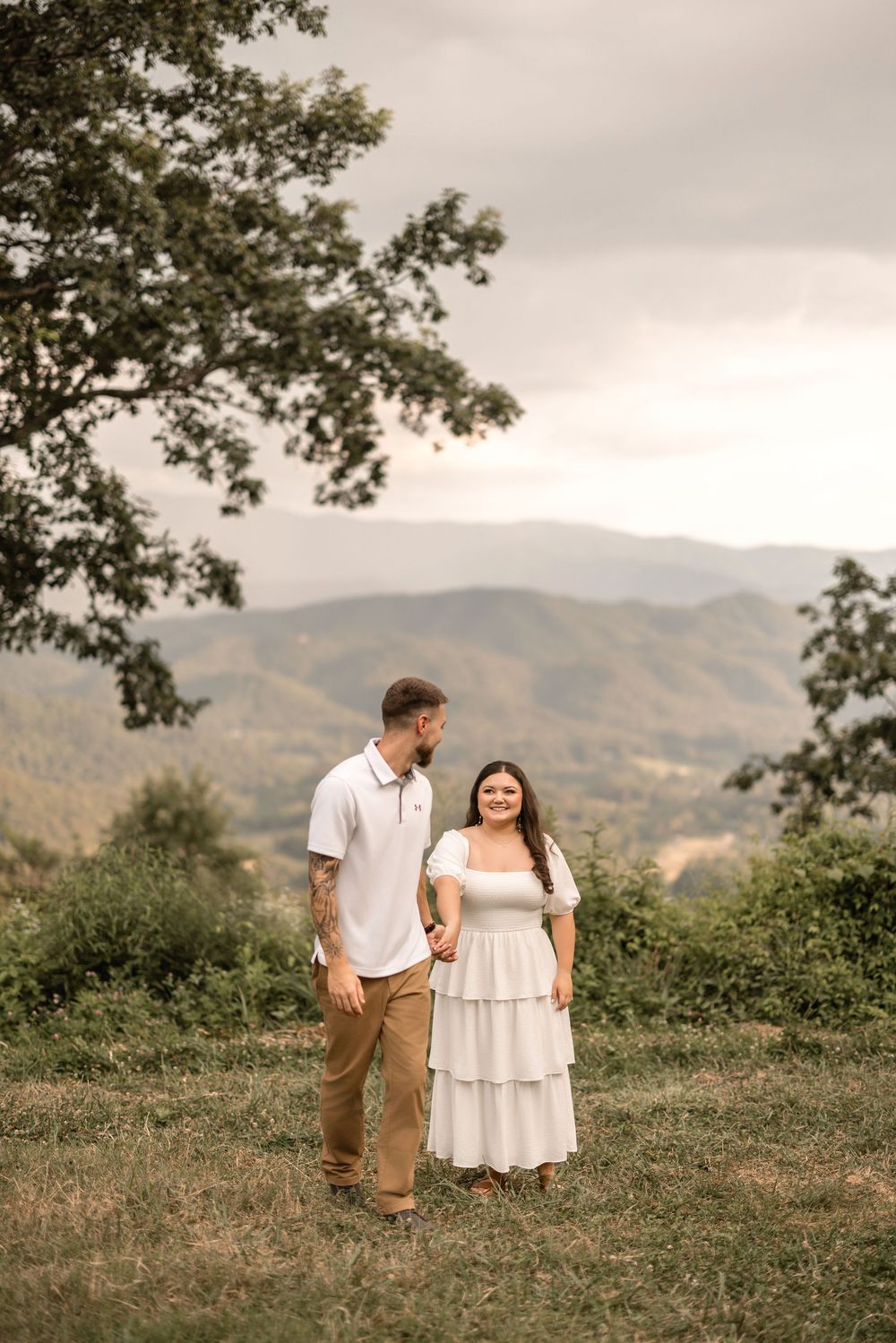 A couple stands together on a grassy overlook with mountain views in the background on a cloudy day.