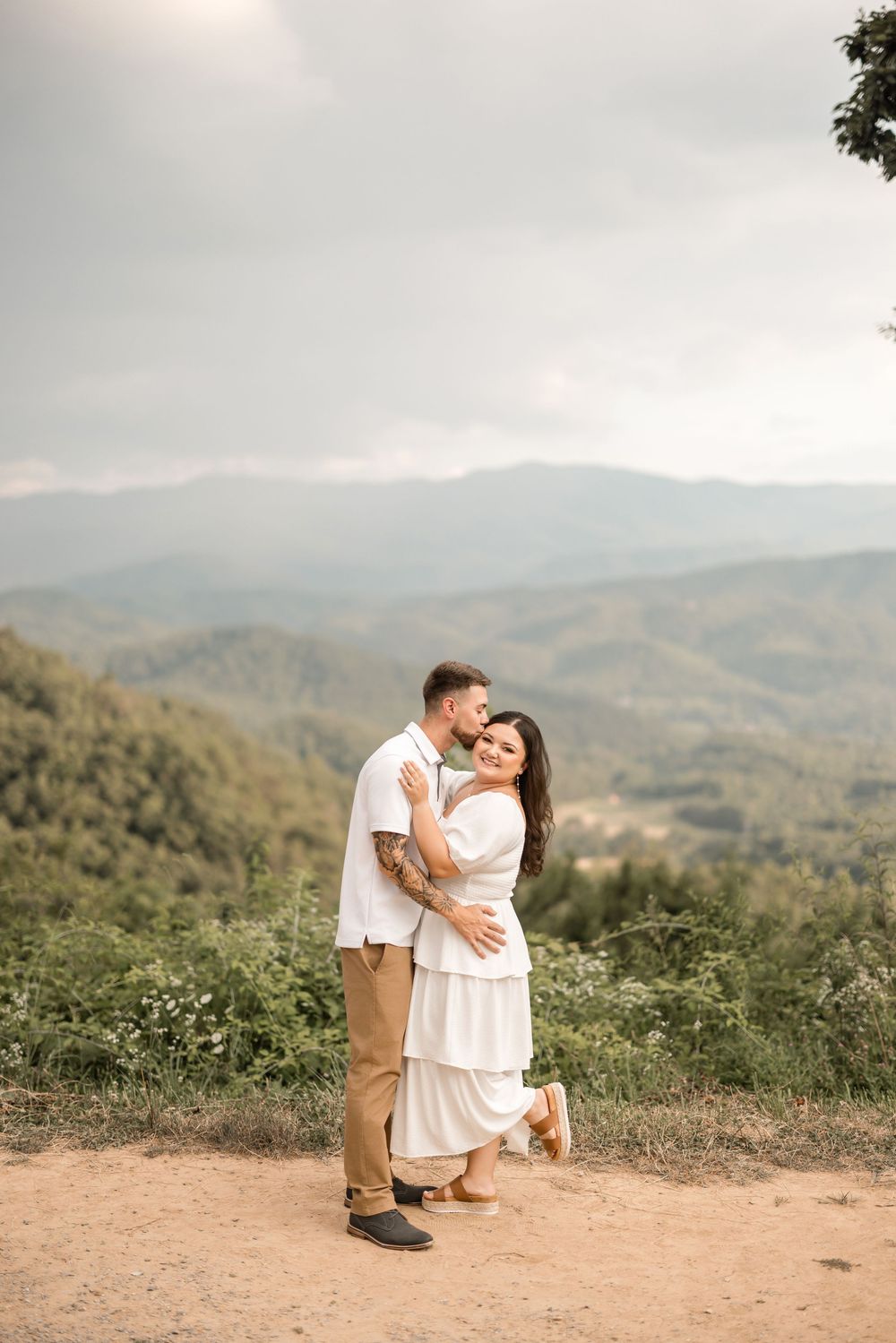 A couple in white outfits share a romantic embrace on a mountaintop overlook with misty rolling hills in the background.