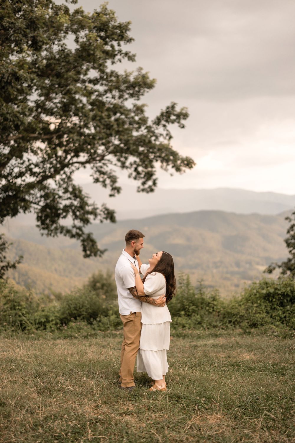A couple stands embracing on a hilltop meadow with mountains and a large tree branch in the background.