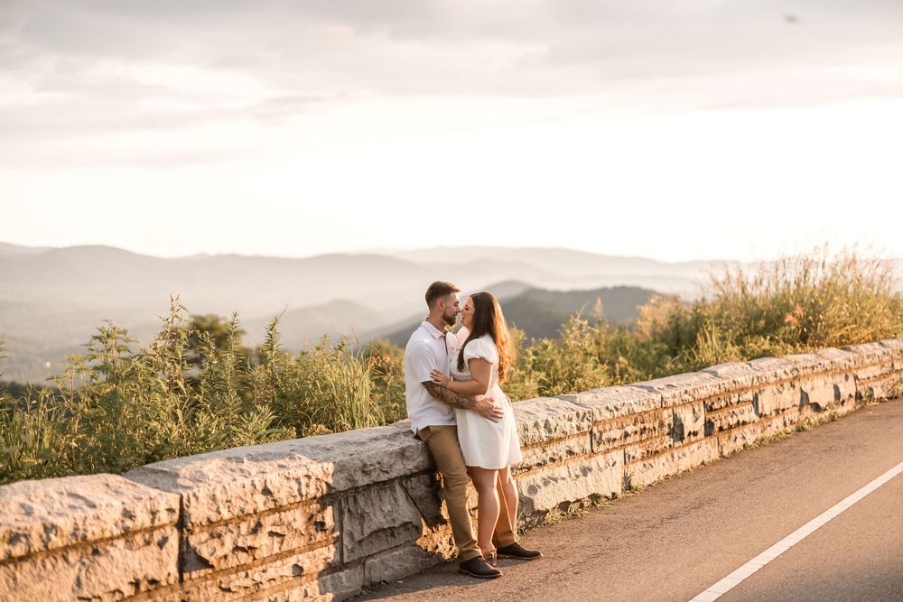 Romantic couple embracing at scenic mountain overlook next to stone wall at sunset.