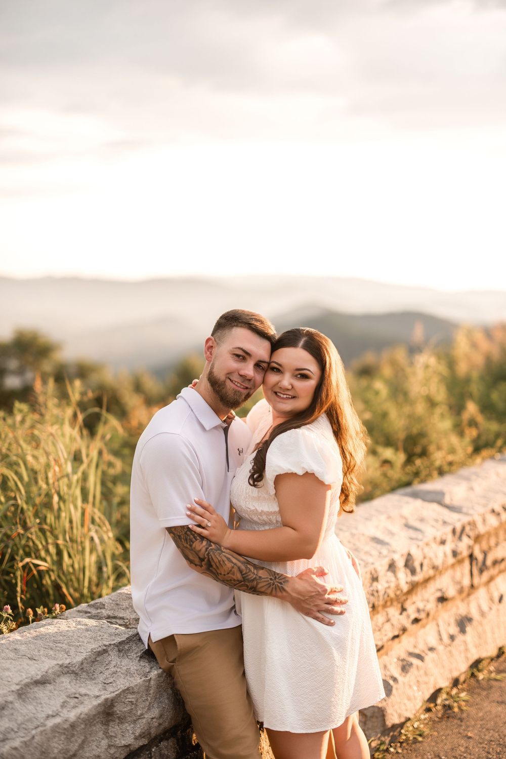 Romantic couple embraces on mountain overlook at sunset with golden grasses and mountains in background.