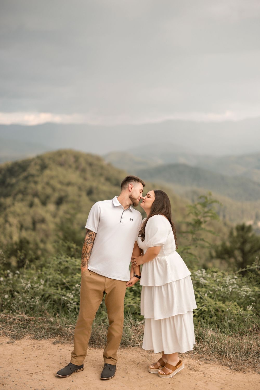 A romantic couple sharing a kiss on a scenic mountaintop overlook surrounded by rolling hills and green foliage at sunset.