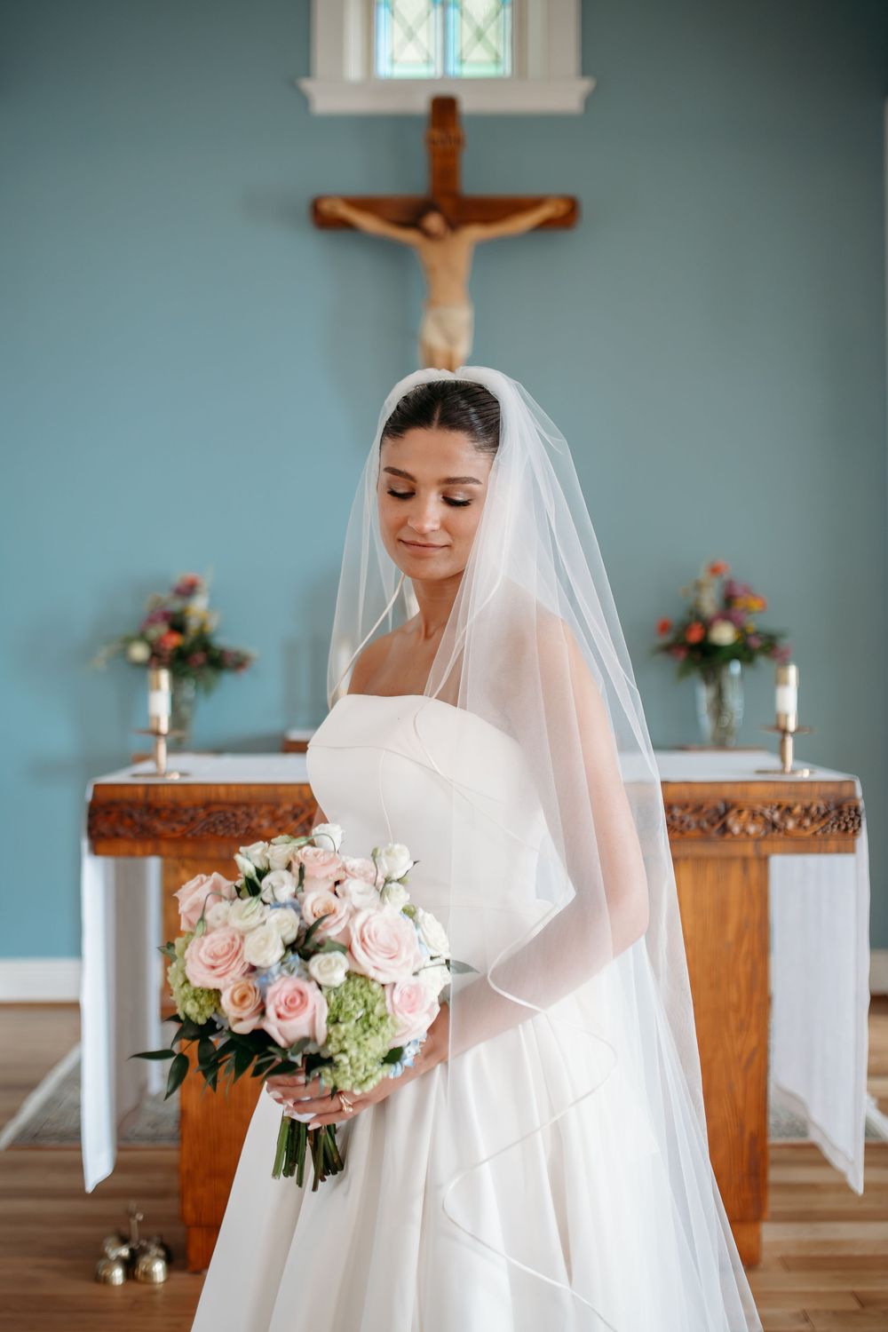 A bride in a white wedding dress and veil stands in a church sanctuary holding a pink and white rose bouquet.
