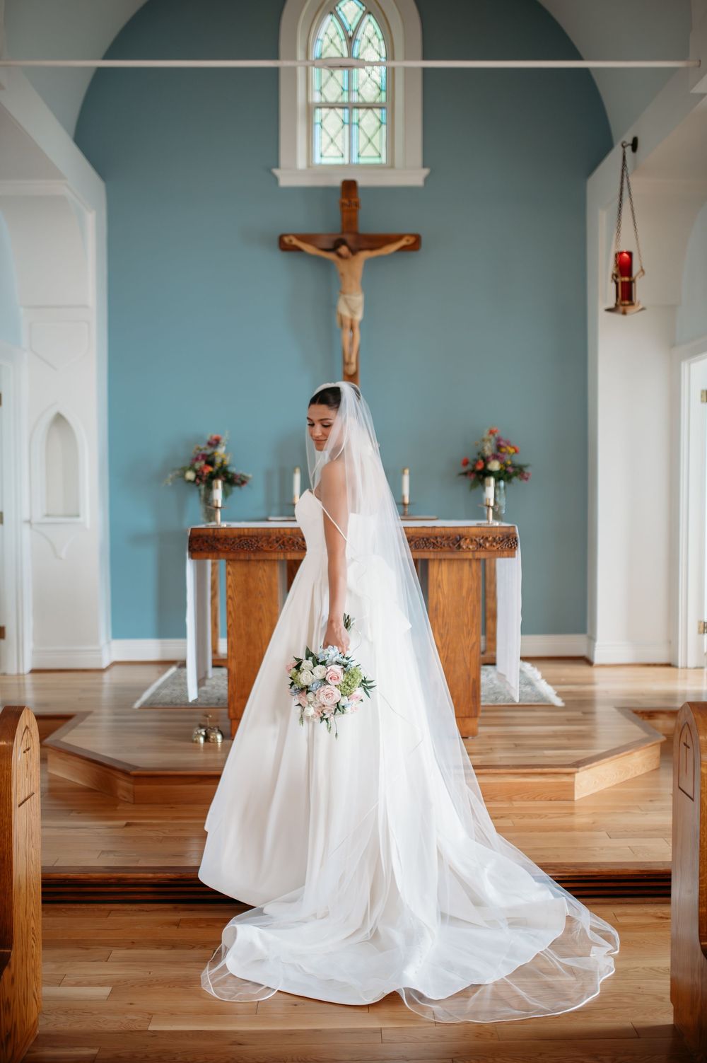 A flowing white wedding dress trailing across the wooden floor of a simple church sanctuary with blue walls and arched windows.