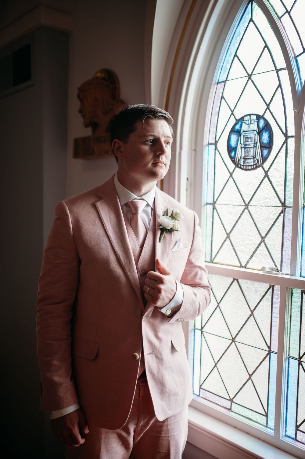 Person in pink suit jacket poses by stained glass window in series of artistic portrait photographs.