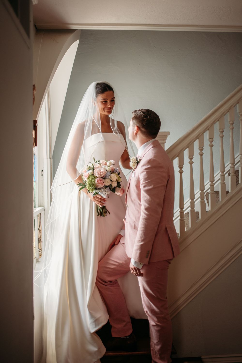 A couple shares a romantic moment on a staircase, with the bride in white and partner wearing a pink suit.