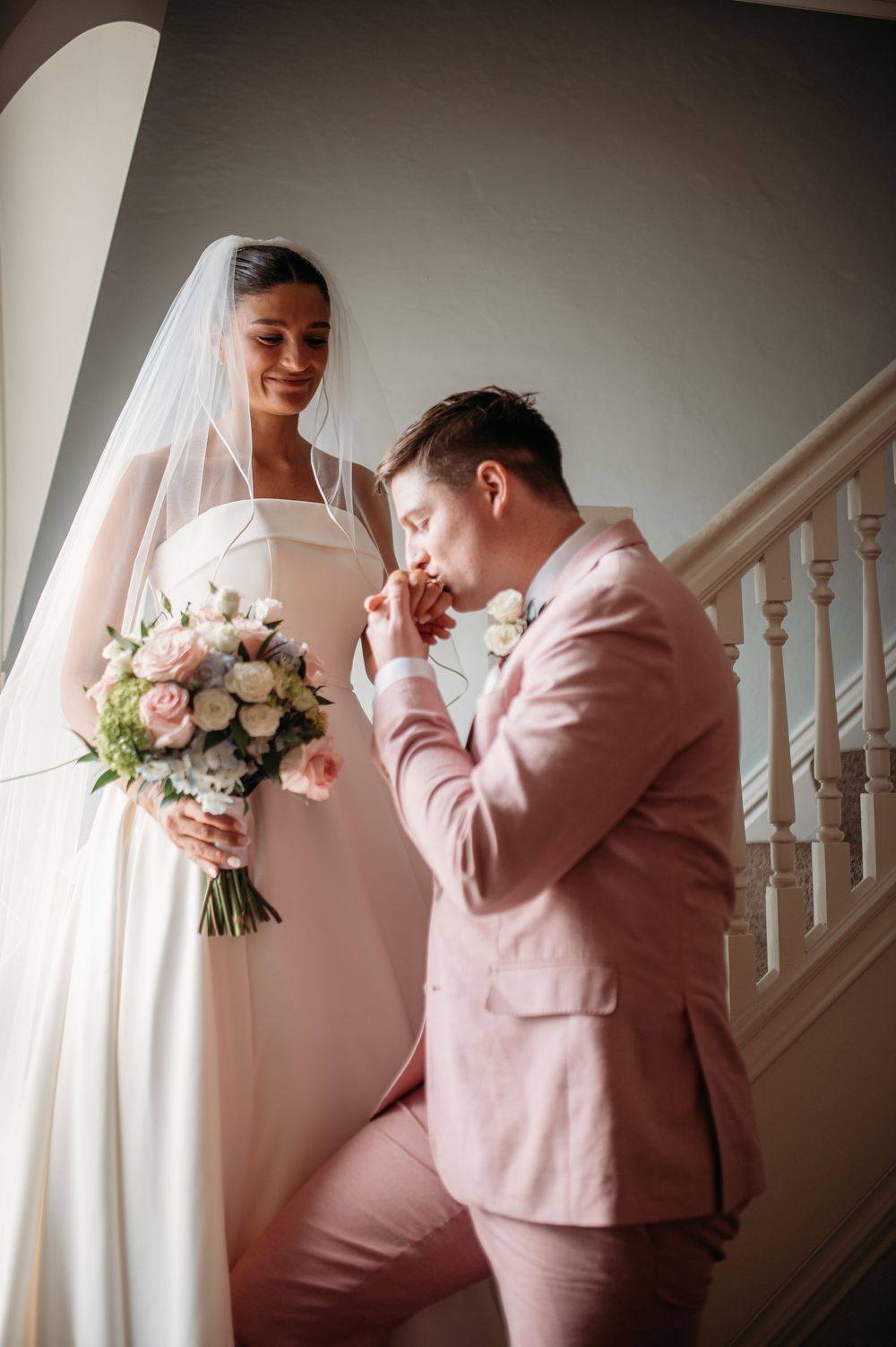 A bride wearing a white wedding dress holds a bouquet while being greeted by a bridesmaid in a pink suit on a staircase.