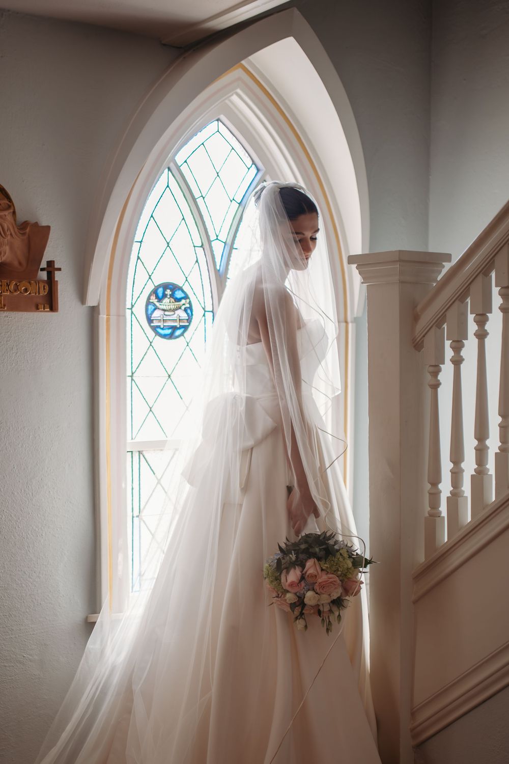 A bride in a flowing white veil and gown stands elegantly by a gothic arched window holding a pink floral bouquet.
