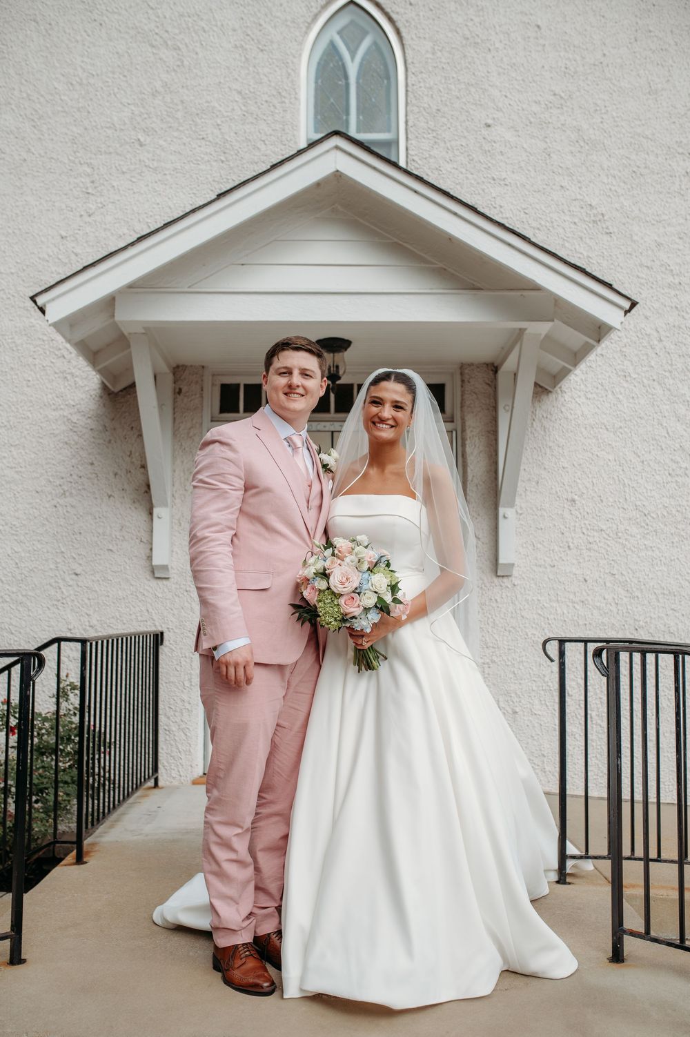 Wedding couple in romantic poses outside a white chapel entrance with black railings, featuring pink and white attire.