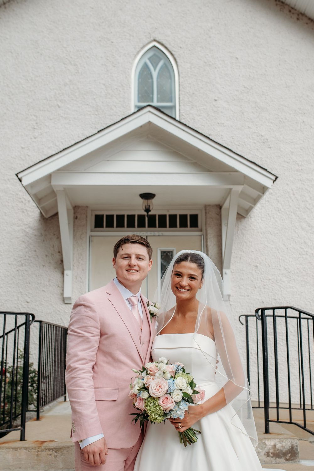 A wedding couple stands in front of a white church entrance with an arched window, groom in pink suit and bride in white dress.