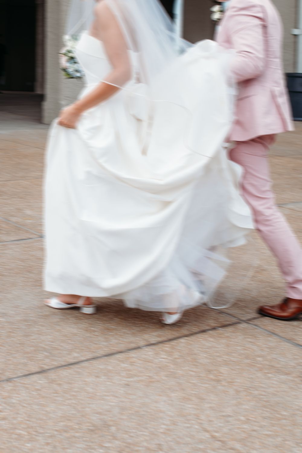 Motion blur shot of a white wedding dress and pink suit walking across a tiled floor.