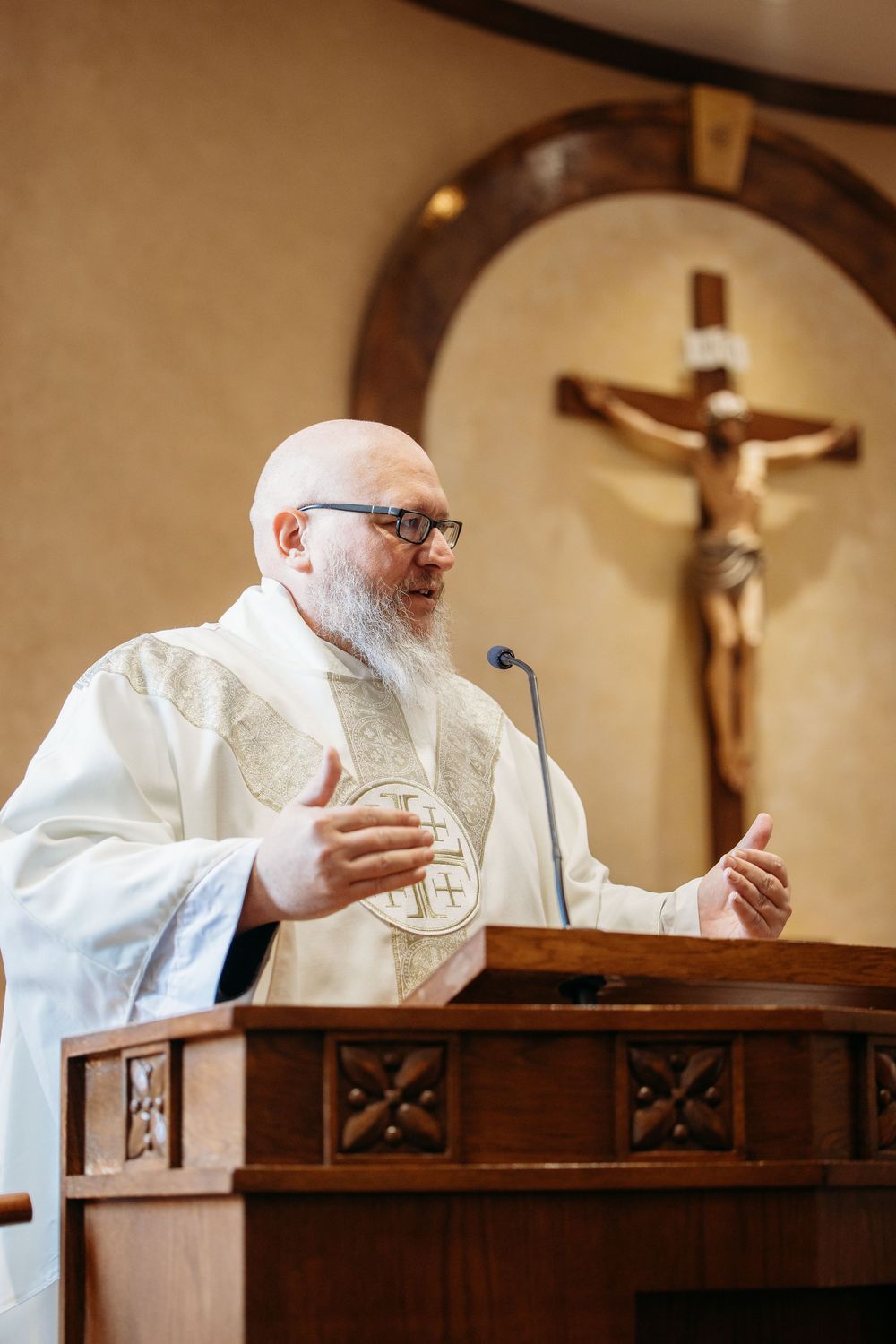 A Catholic Priest in white robes gestures while giving sermon at wooden pulpit with crucifix visible in church background in St. Louis, MO.