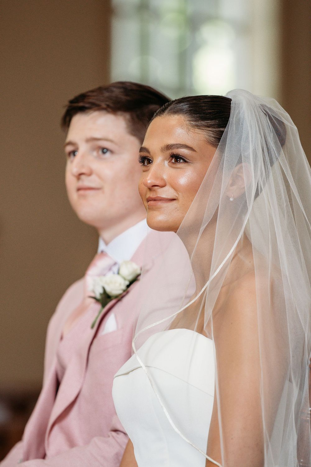 A bride in a white dress with veil sits next to her partner wearing a pink suit during their wedding ceremony.