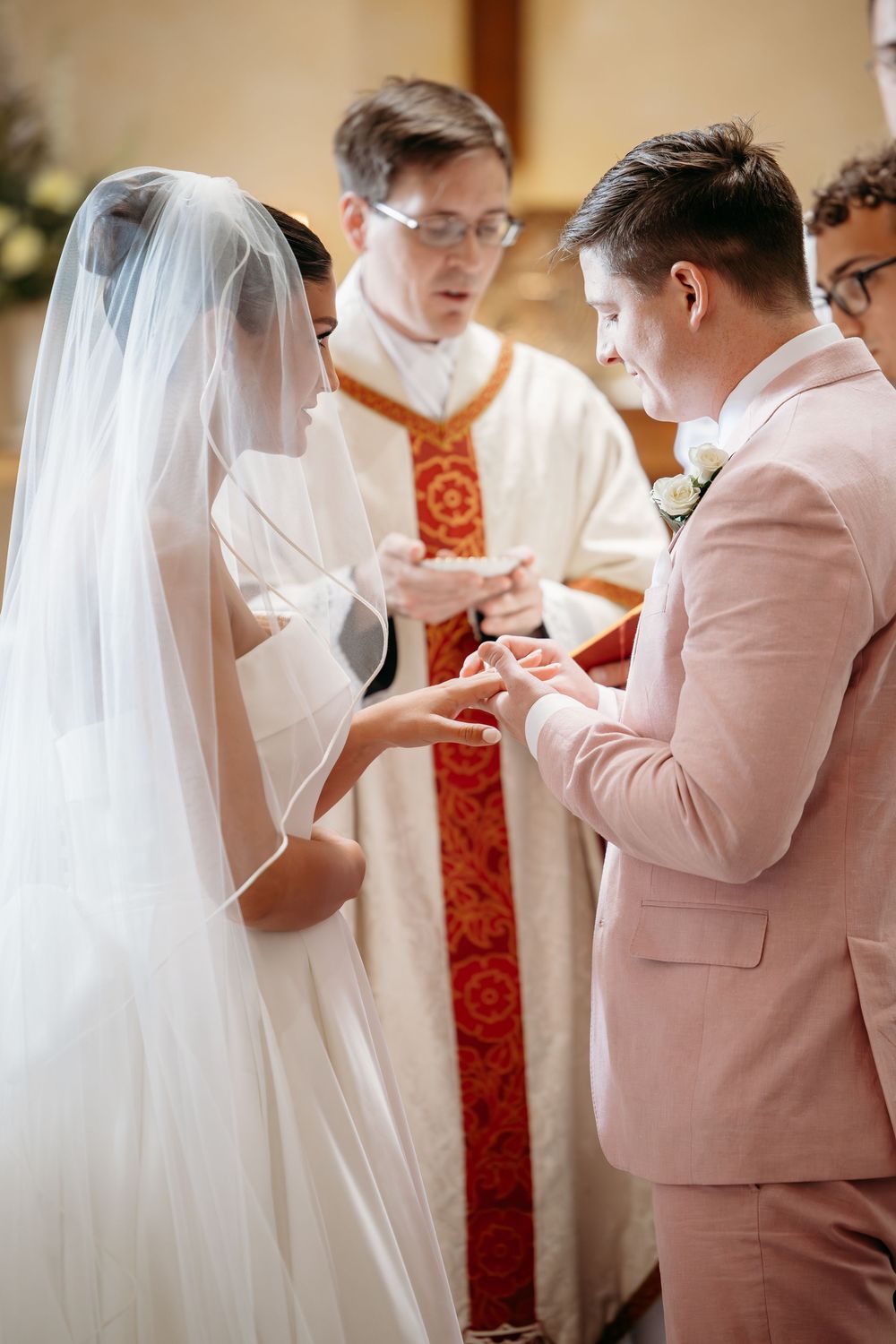A couple exchanges rings during a wedding ceremony as an officiant in red and white vestments presides.