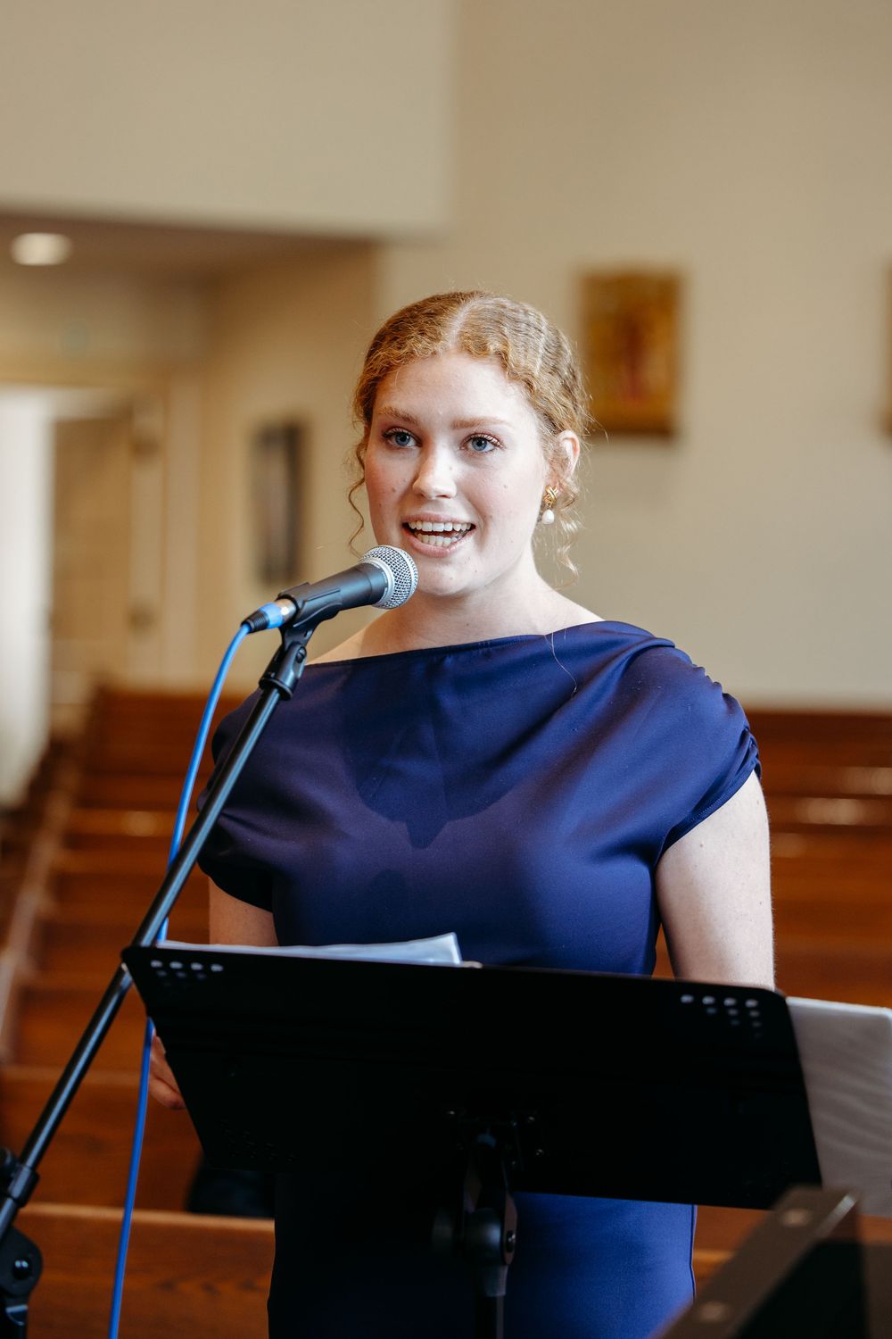 Speaker wearing navy blue dress stands at microphone with sheet music stand in church setting.