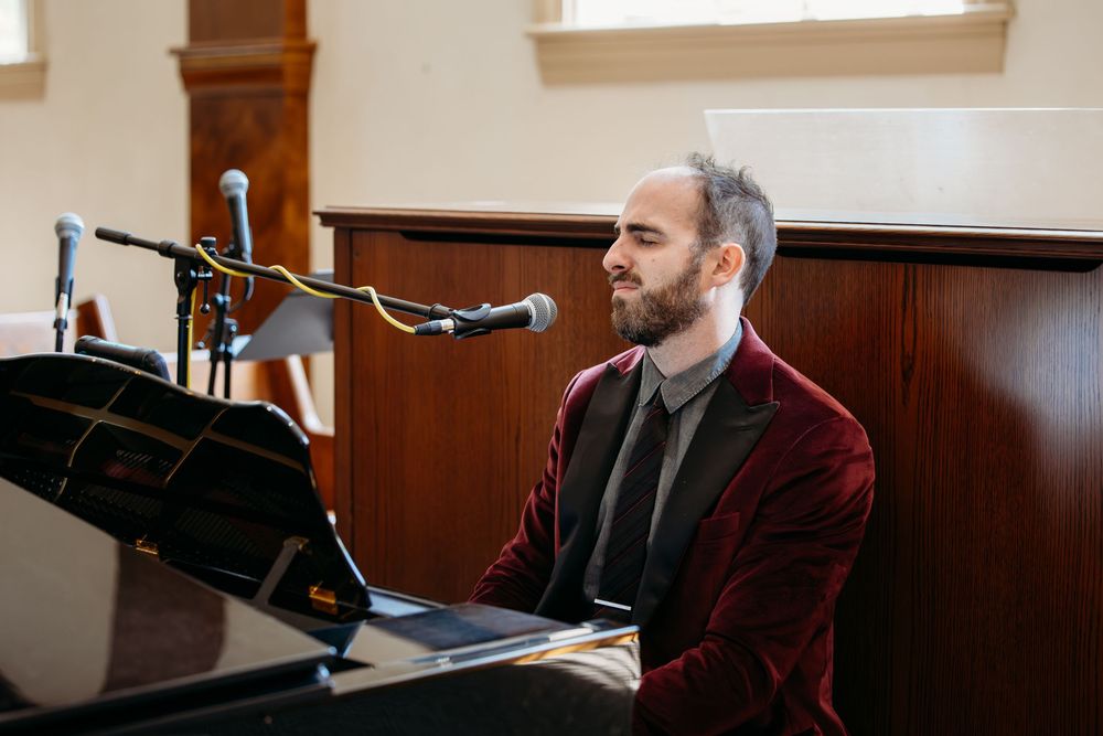 A pianist in a burgundy blazer sits at a grand piano with a microphone stand beside them during a performance.