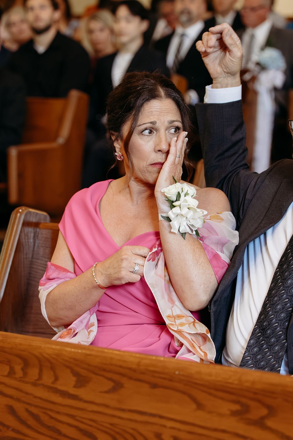 An emotional wedding guest in a pink dress and white corsage wiping tears and raising a hand during the ceremony.