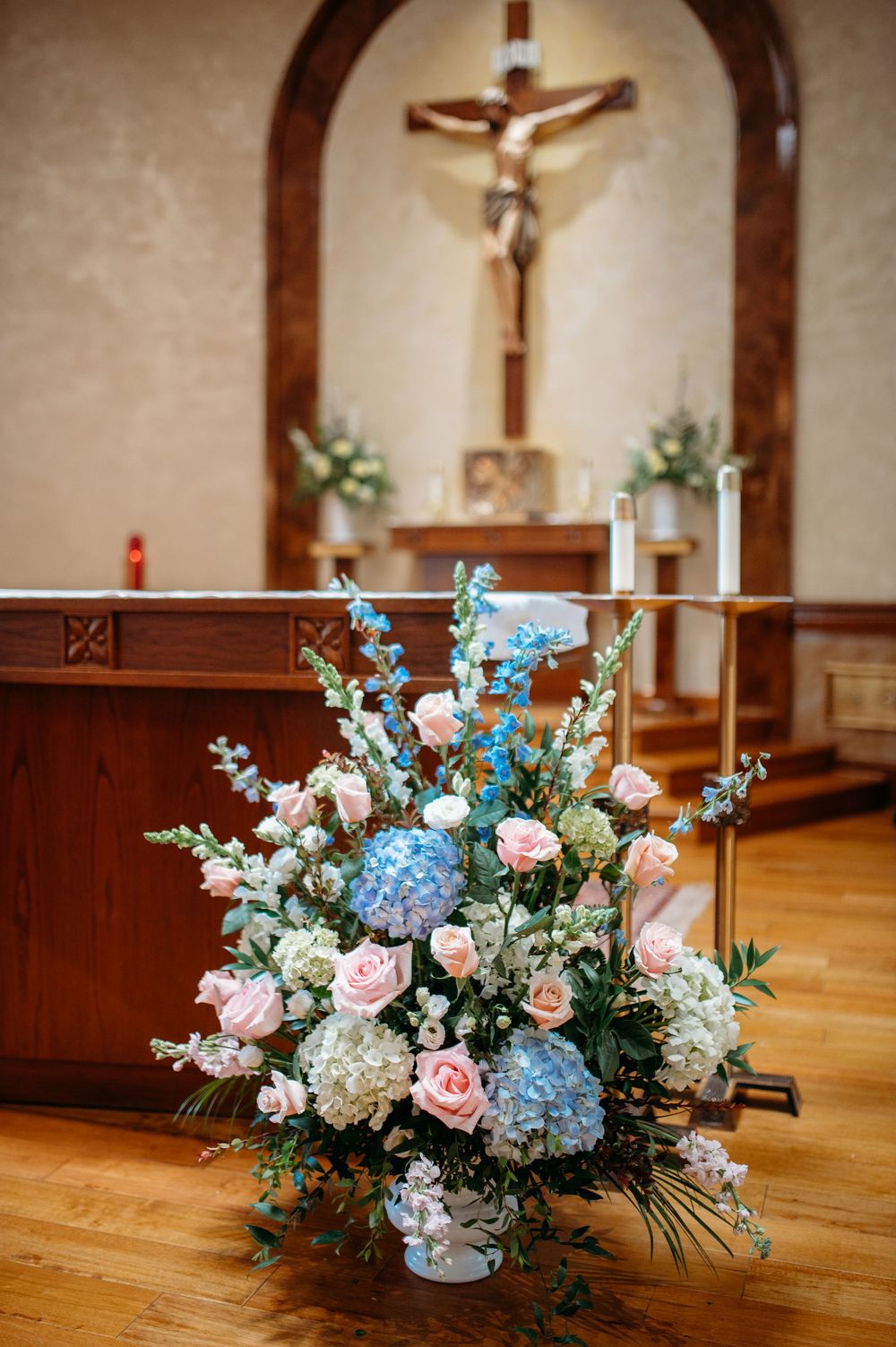 A church interior showcasing blue hydrangeas and pink roses in floral arrangements with a wooden crucifix in the background.
