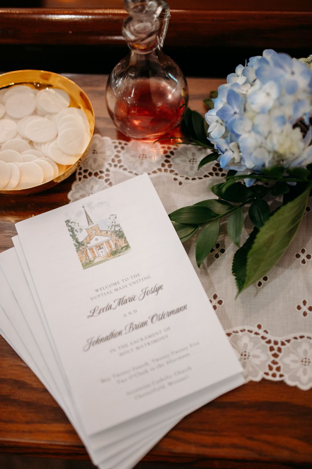 Wedding invitations with a watercolor house illustration displayed with blue hydrangeas and crystal decanter on lace tablecloth.