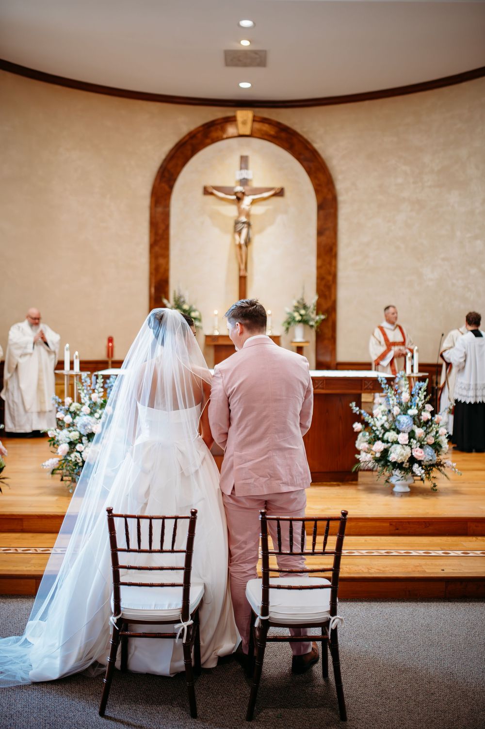 A couple sits on wooden chairs during their Catholic wedding ceremony in a church adorned with floral arrangements.