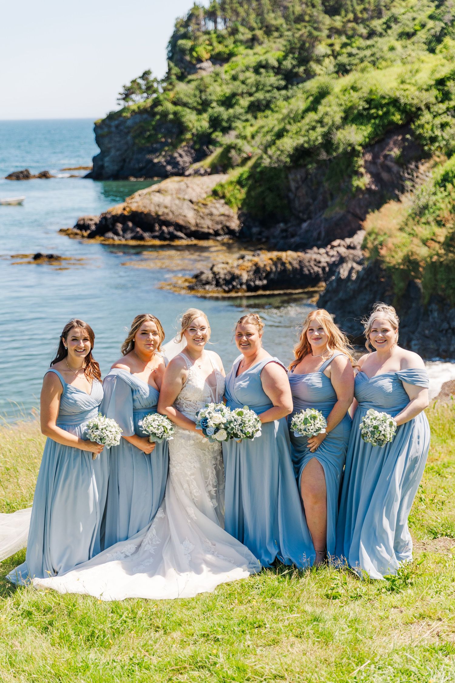 A wedding party poses on a grassy cliff overlooking a scenic rocky coastline on a bright sunny day.