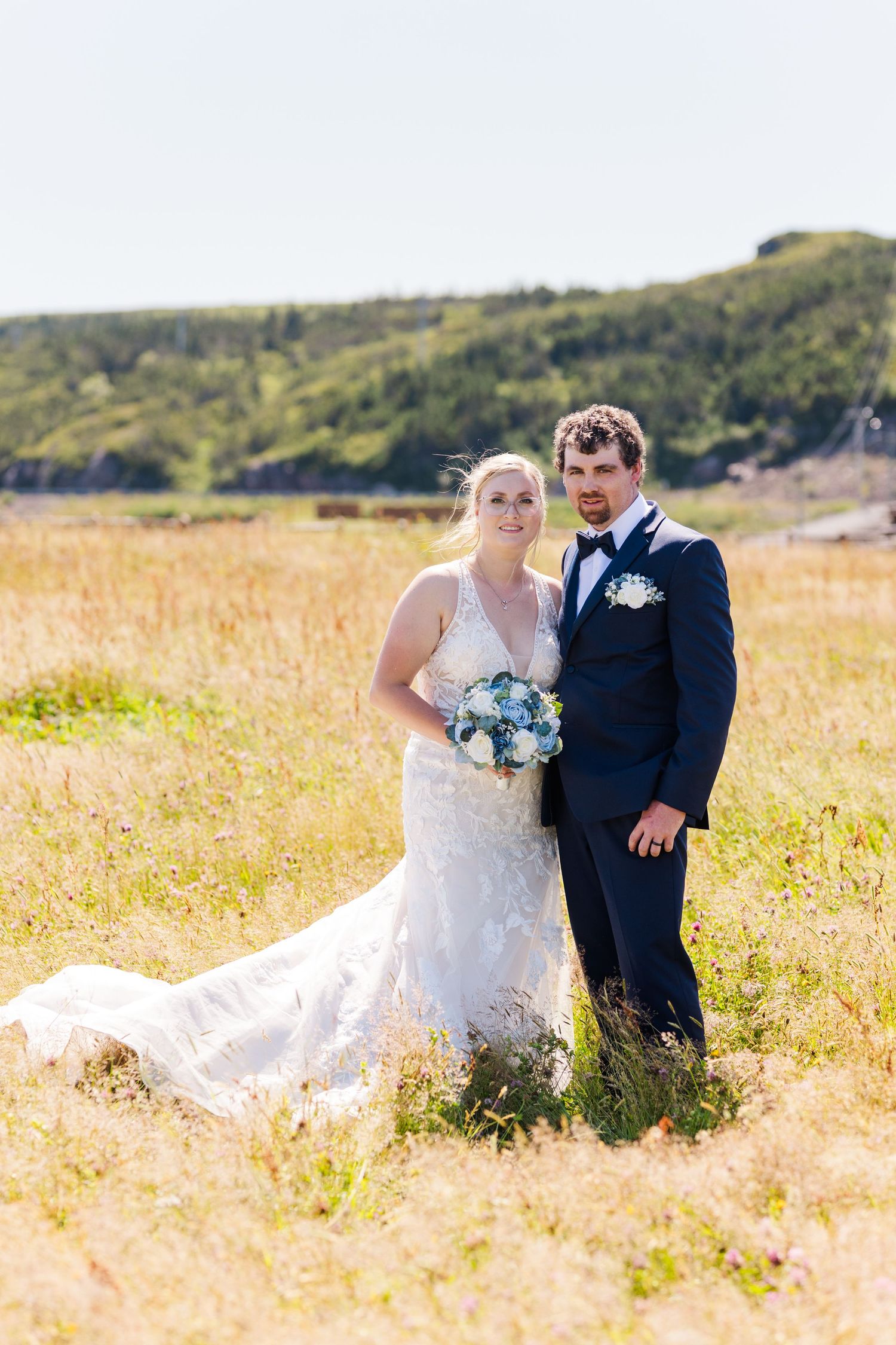 Wedding couple stands in a sunlit meadow with hills in background and blue bouquet against navy suit and white dress.