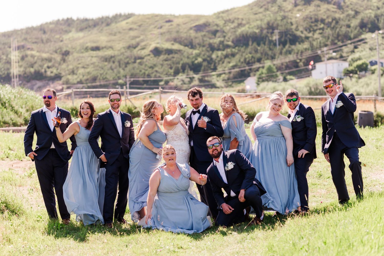 Wedding party in navy suits and light blue bridesmaid dresses pose together on grassy hillside.