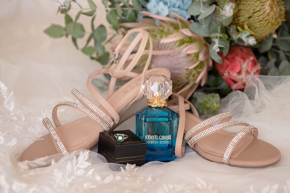 Wedding details featuring nude strappy sandals, perfume bottle, and floral arrangements on white lace background.