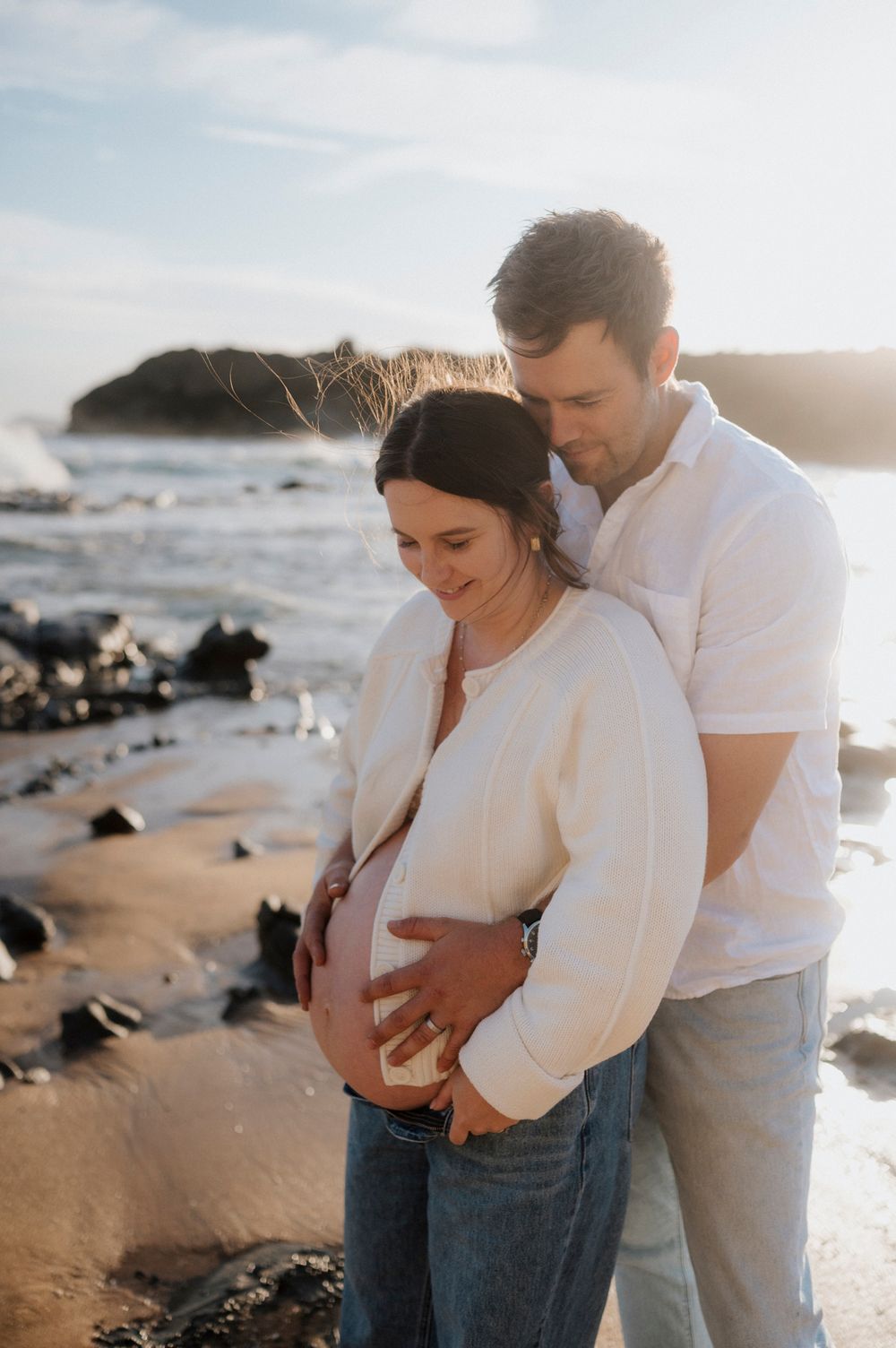 A couple embraces tenderly on a rocky beach at sunset with waves crashing in the background.