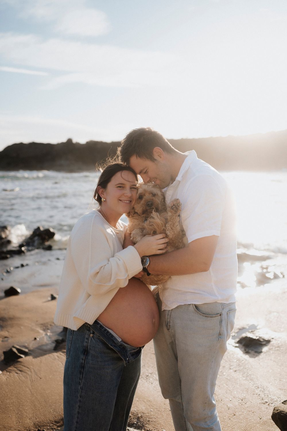 Couple embraces with small dog on beach at sunset, with expectant mother showing pregnant belly in casual attire.