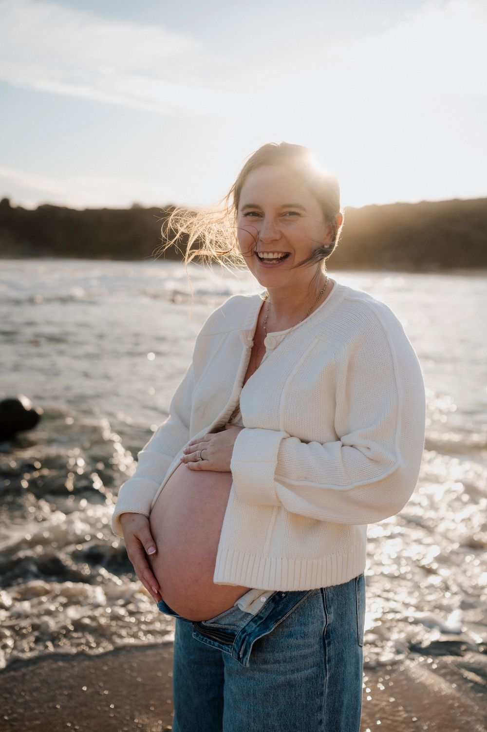 A pregnant woman in a white sweater smiles joyfully while standing on a rocky beach at sunset.